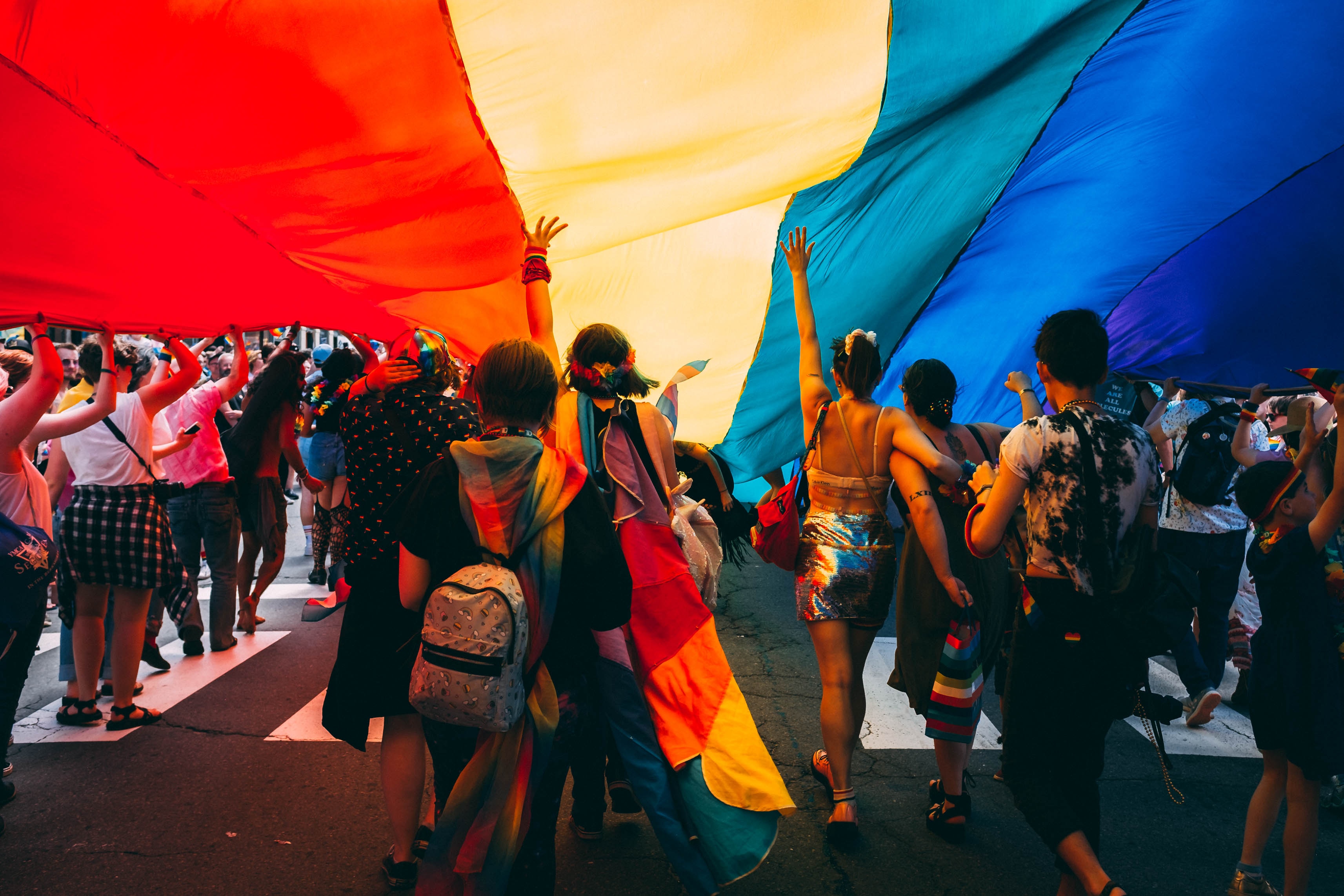 large group of people at a pride festival under a huge rainbow flag that they are walking under 
