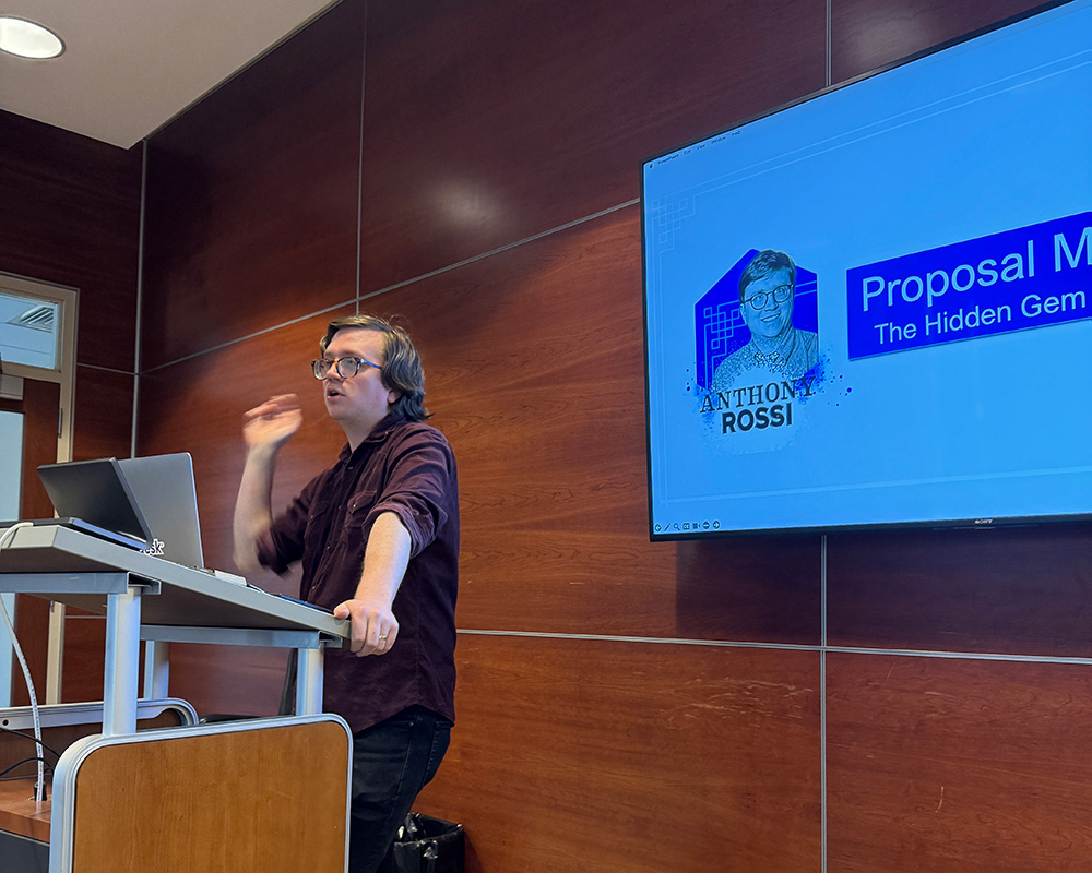 Anthony standing at a podium with a tv with his presentation in the background