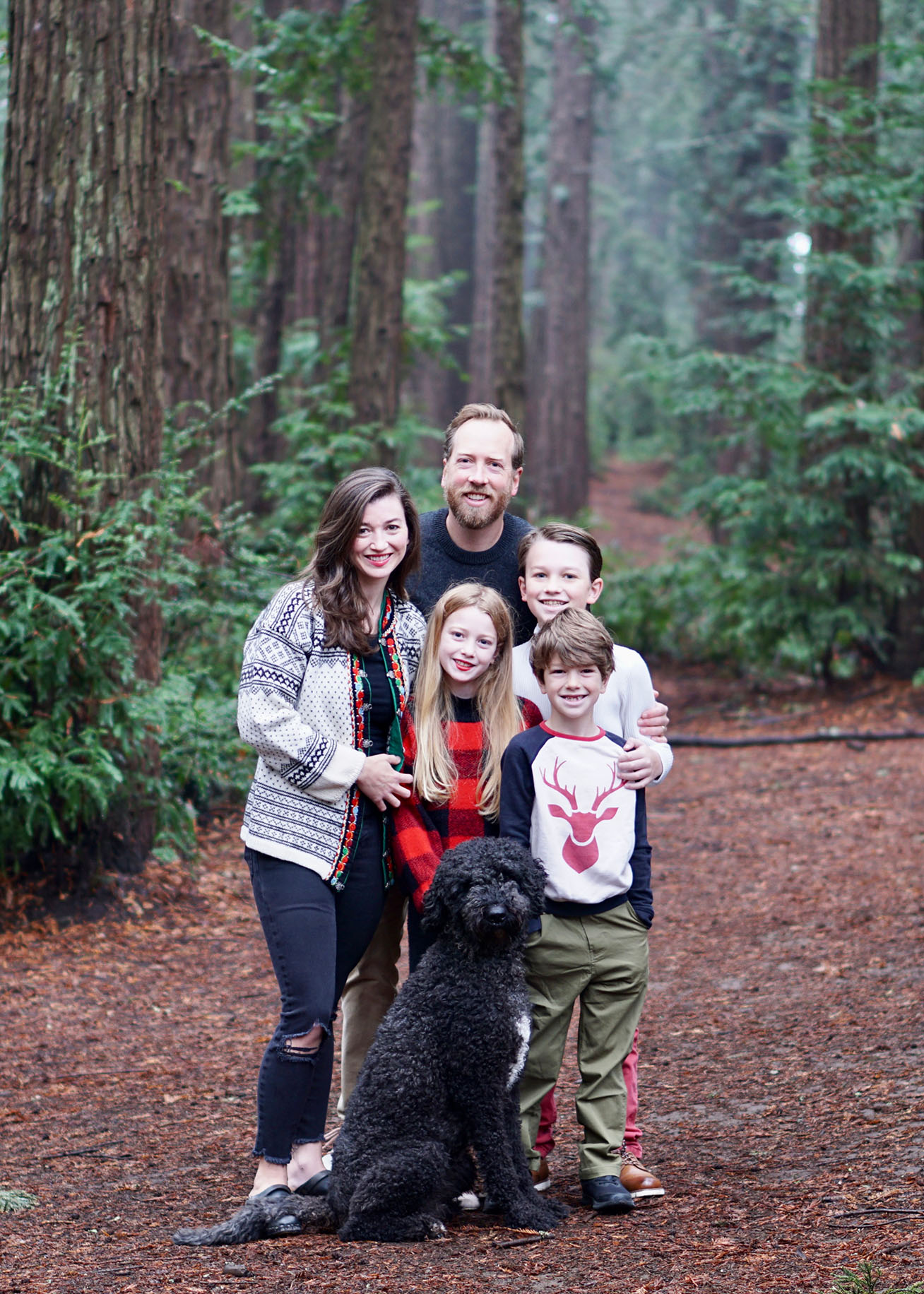 Kevin Knight and his family of 5 stand in a forest with large pine trees. 