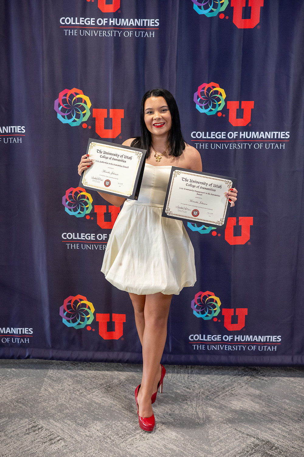 A student in a white dress holds awards in front of the College of Humanities photo backdrop