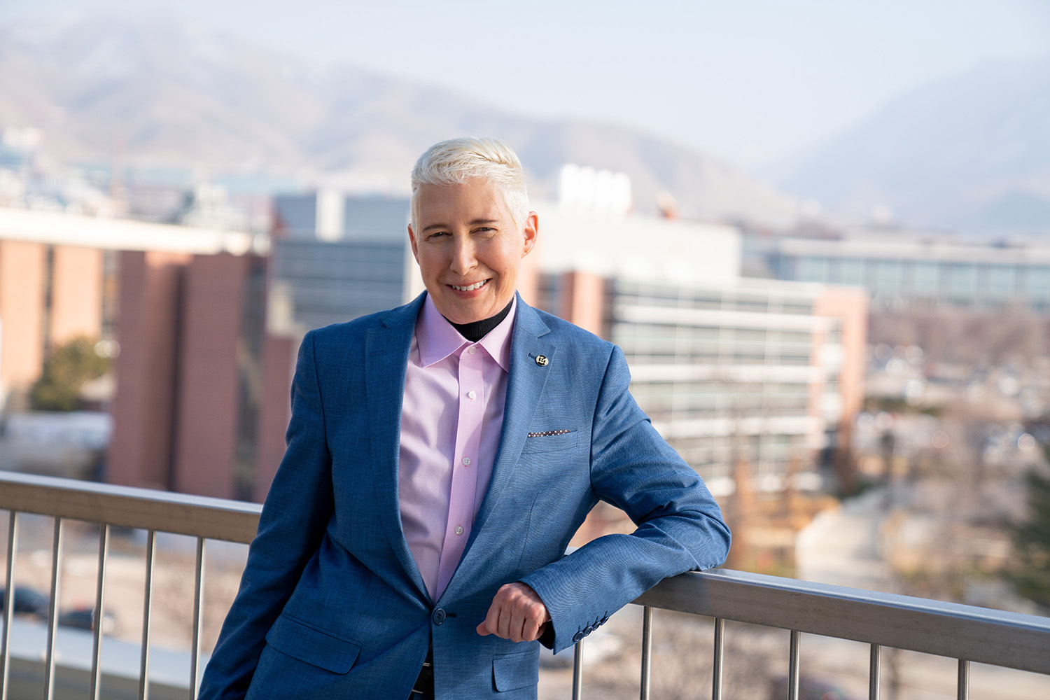 Kathryn Bond Stockton leans on a balcony railing with a background of the Wasatch mountains and U of U campus