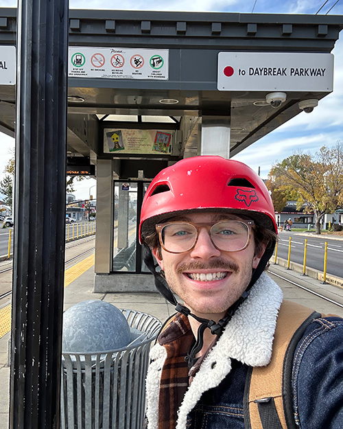 Kellen wearing a red helmet and glasses smiling at the camera standing at a trax station.