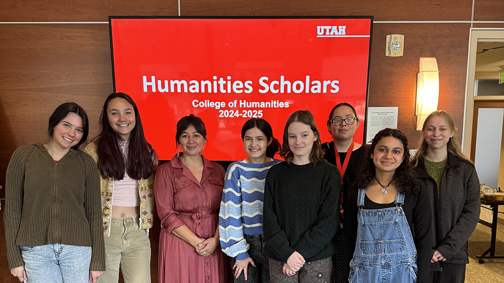 7 young women stand together in a line as a group. They are all smiling at the camera with a tv showing a red background with 'Humanities Scholars' on the screen
