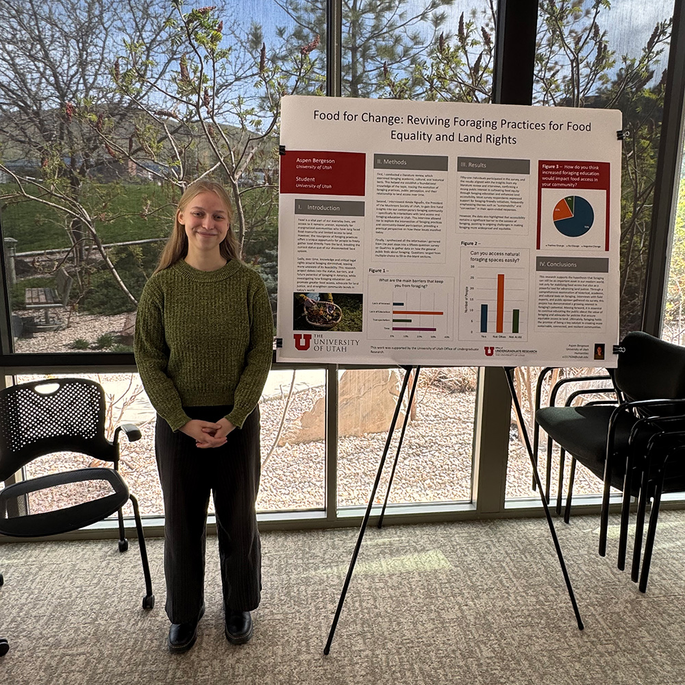 A young woman with long blonde hair stands with her hands crossed in front of her. Her reseach poster is the the right-side of her.