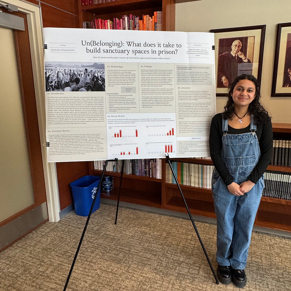A young woman with long dark hair stands with her hands crossed in front of her. Her reseach poster is the the left-side of her.