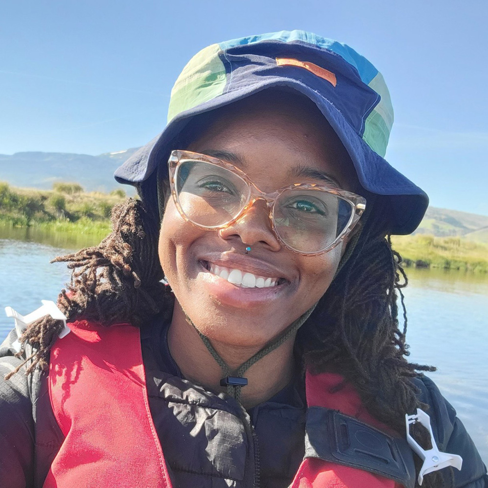 A young woman smiling at a camera. She is wearing a lifevest and a bucket hat with a body of water in the background