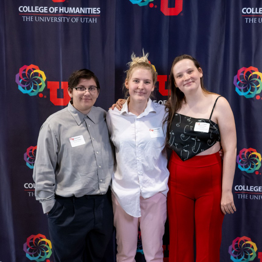Three young people stand in front of a purple banner with College of Humanities branding on it.
