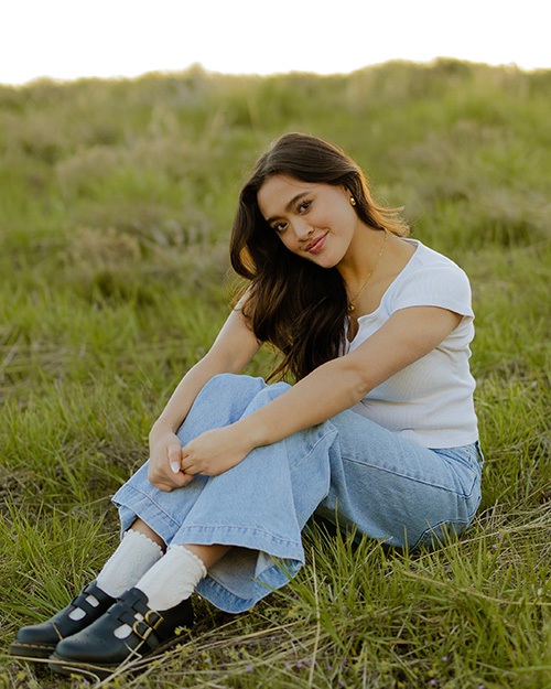 Amy sitting on a grass hill smiling at the camera. She is wearing white blouse and light-colored jeans