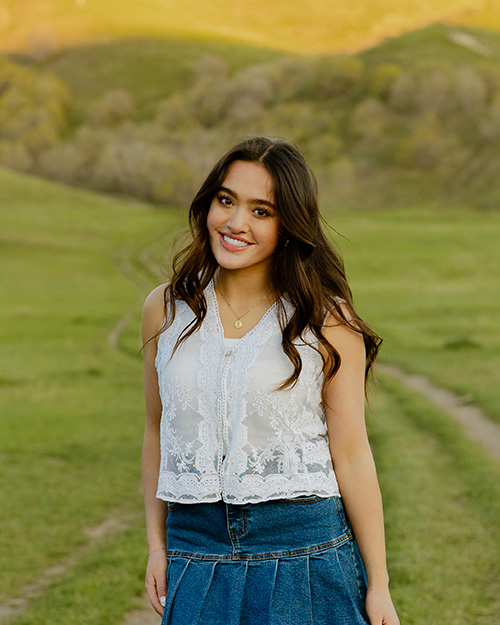 Amy standing in a green field. Her long dark hair is tumbling in front of her has she smiles at the camera. She is wearing a denim skirt and white blouse