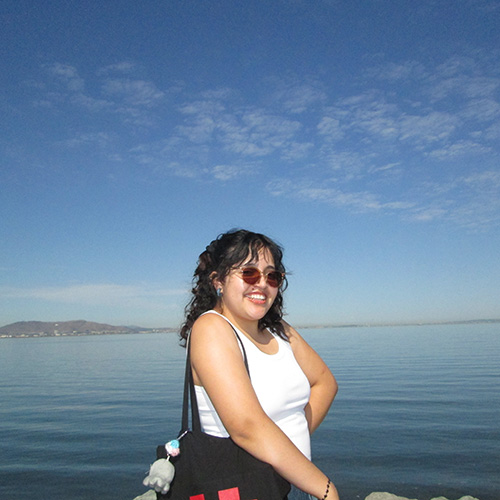 Brittney is standing in front of a calm lake and clear blue sky. She is wearing sunglasses and smiling at the camera