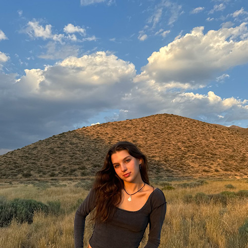 Katherine is wearing a long sleeve black top. There is a grass field, mountains, and blue sky with fluffy white clouds in the background.