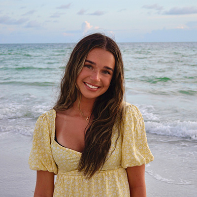 Katie is standing smiling at the camera. She is wearing a yellow dress and has a blue water beach with soft waves in the background