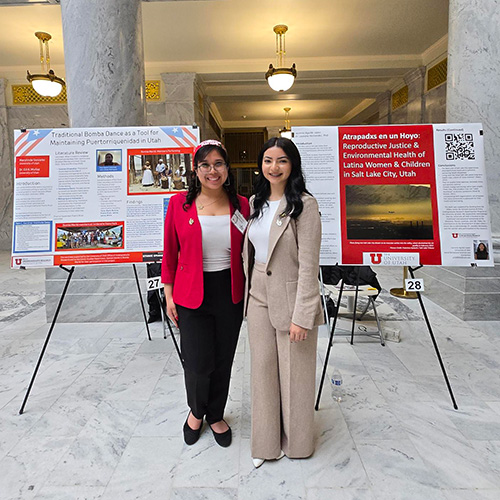 Marylinda and Jasmine wearing business wear standing in front of their research posters with marble pillars behind them. 