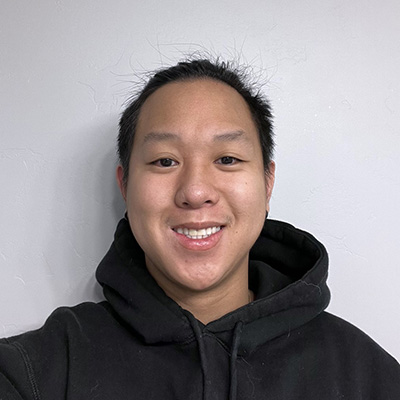 Nathan standing against a white wall. He is smiling at a camera and wearing a black hoodie