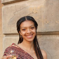 Nayra has long braided hair. She is standing in front of a stone wall smiling at the camera