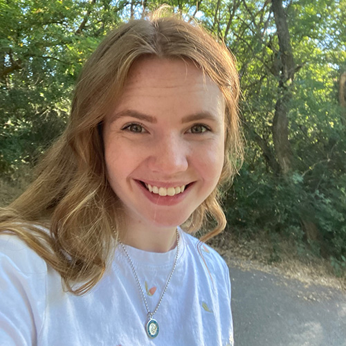 Sophie has shoulder length, wavy, blonde hair. She is smiling at the camera with greenery in the background.