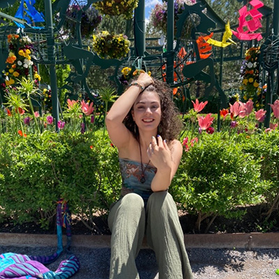 Tara is sitting on the ground with a garden in the background. She has one of her hands in her dark curly hair. The other hand is resting on her knee.