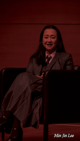 Min sitting on the stage at her lecture. A maroon filter is covering the whole image.